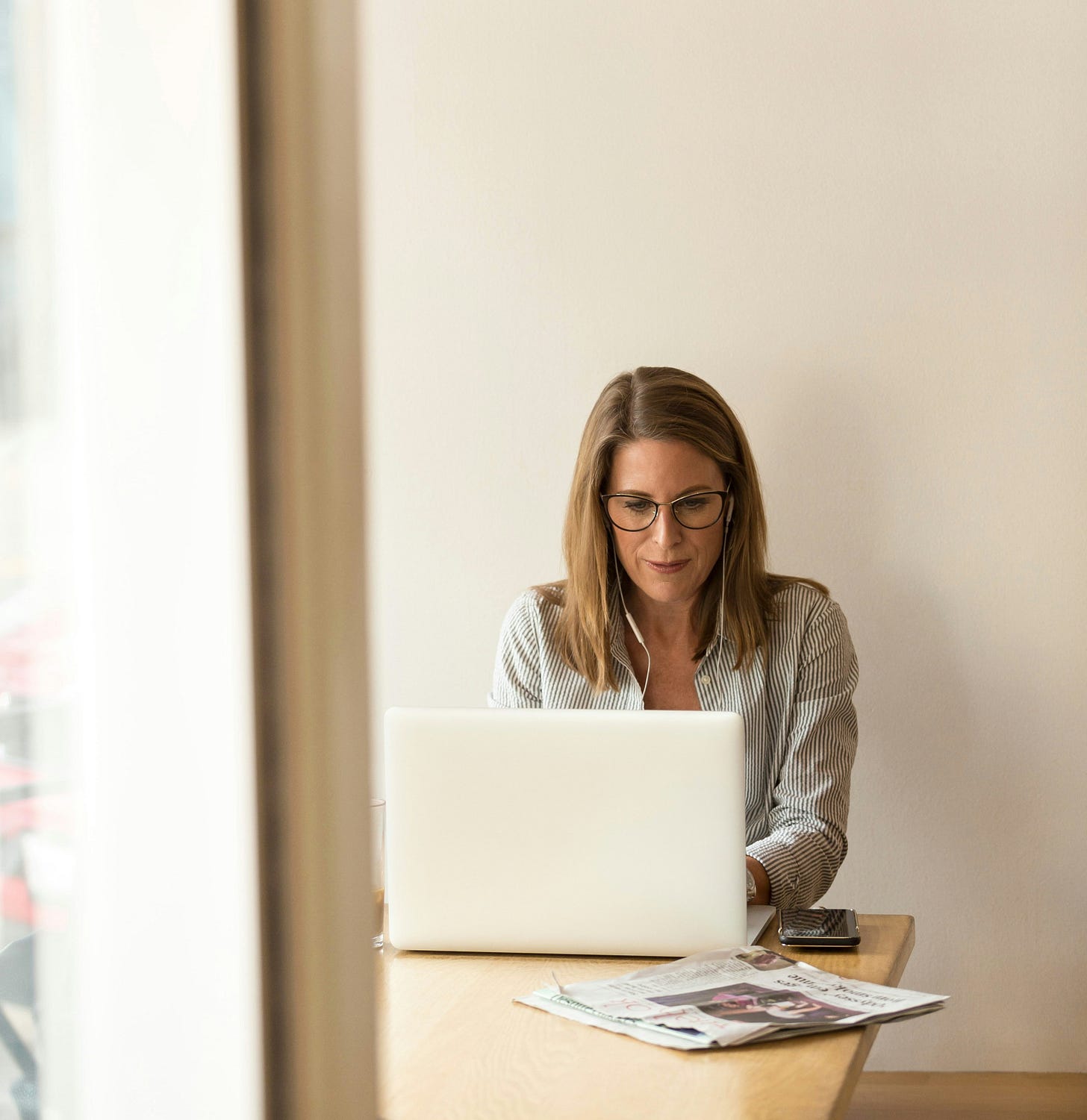 A white woman sits at a table, working on her laptop with white wired headphones in her ears.