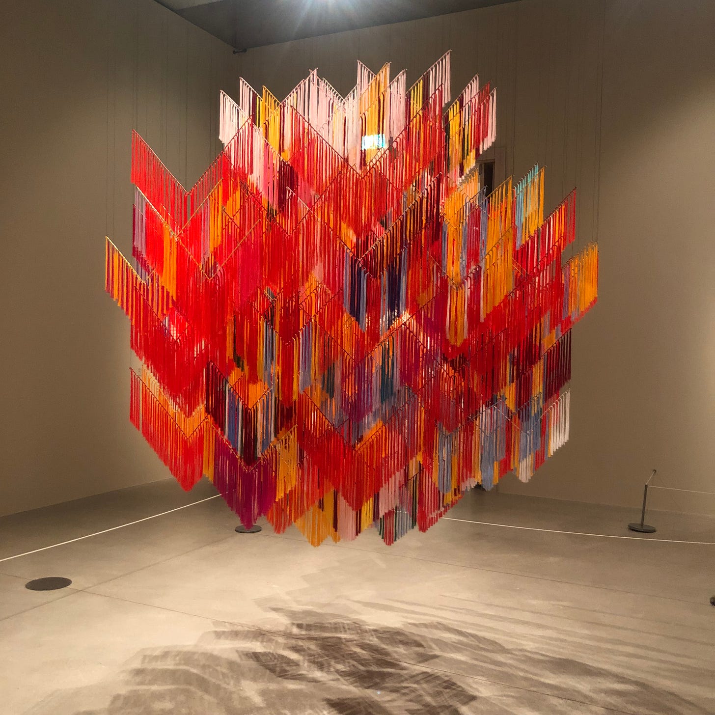 A brightly coloured sculpture made from tied red, orange and purple threads hangs in a gallery space casting shadows onto the floor.