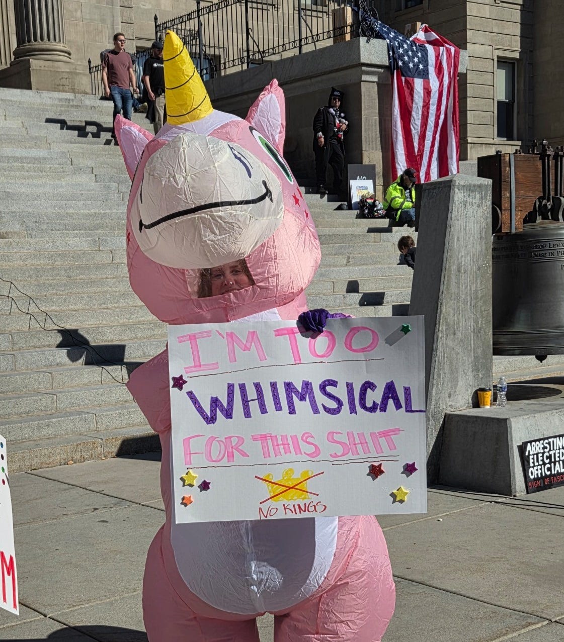 Woman in an inflatable pink unicorn costume in front of the steps of Idaho's Capitol. She carries a  sign reading 'I'm too whimsical for this shit'