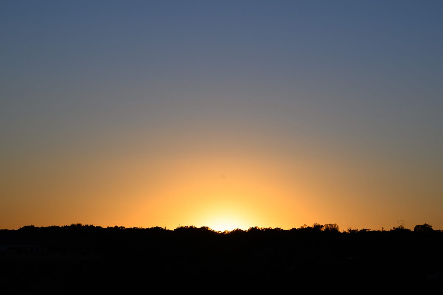 The setting sun above a treeline with a clear blue sky above and the sun’s rays forming a semi-circle above the horizon