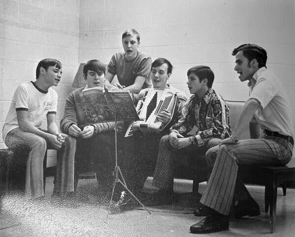 A black and white photograph of six teenage boys sitting and singing. One of them is playing an accordion and looking at sheet music on a stand in front of him.