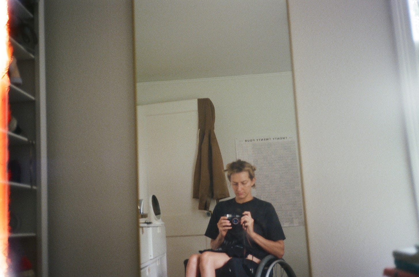 Rebekah, looking down and sitting in her wheelchair, points a 35mm camera at a long vertical mirror. Behind her, there is a brown jacket hanging on the side of an open door and a giant wall calendar. The left edge of the photo is burned bright orange. Rebekah, looking down and sitting in her wheelchair, points a 35mm camera at a long vertical mirror. Behind her, there is a brown jacket hanging on the side of an open door and a giant wall calendar. The left edge of the photo is burned bright orange.