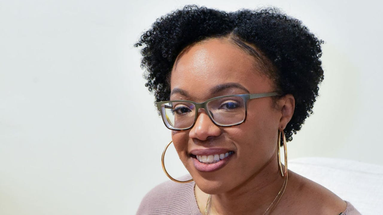 Smiling portrait of Tomesha Campbell wearing glasses and gold hoop earrings, with natural curly hair styled in two puffs, against a light background.