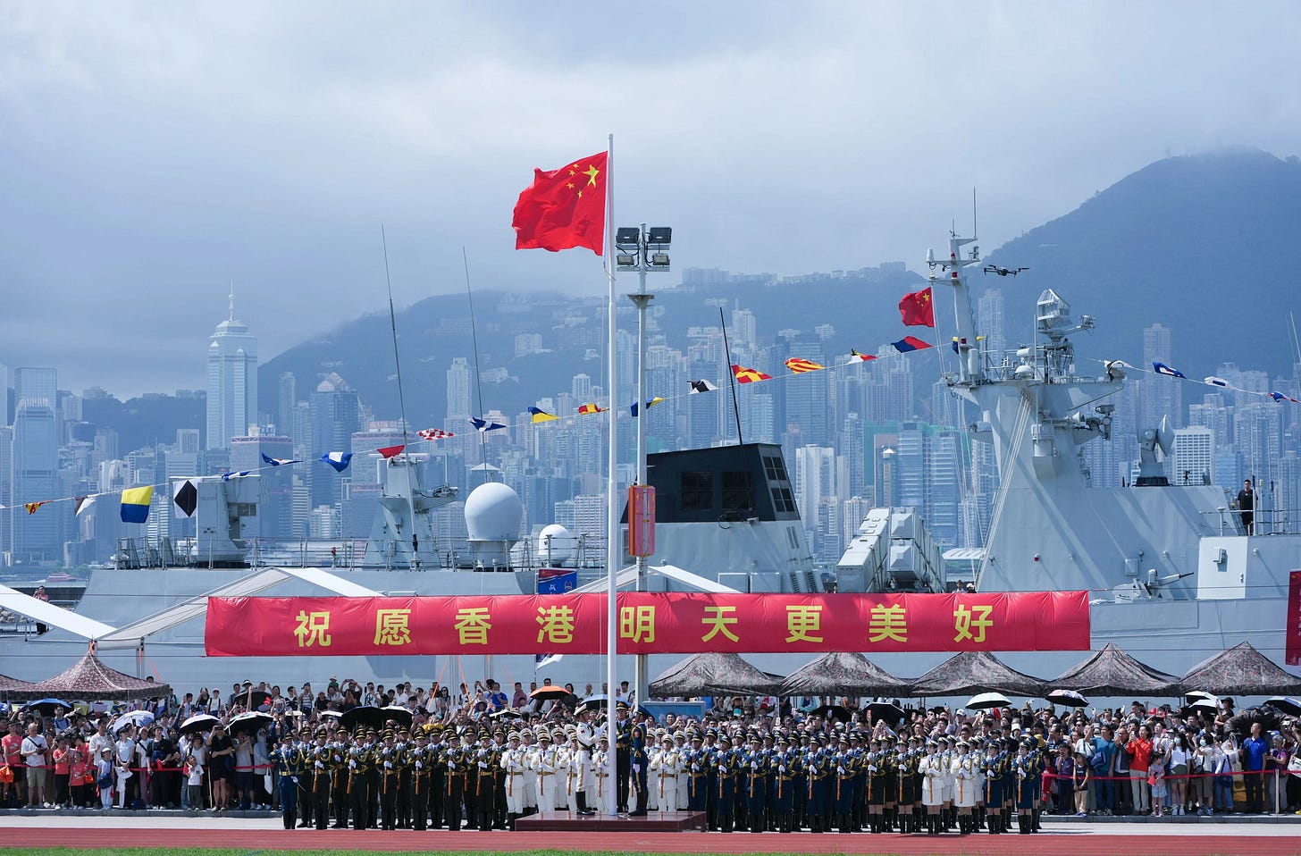 PLA Hong Kong Garrison public open day activities (June 20–28, 2026) — Crowds visiting barracks with military formations, Chinese flags, and naval vessels in the background.