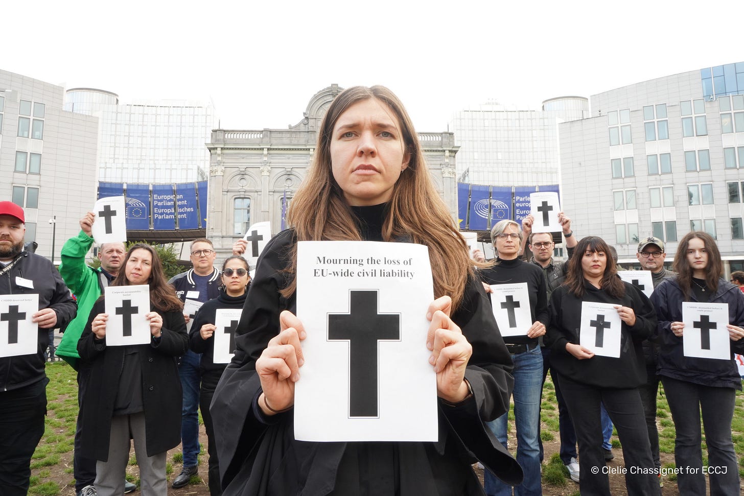 A group of people stand in Place Luxembourg in front of the European Parliament dressed in black and holding pieces of paper with crosses on them and the text 'Mourning the death of EU-wide civil liability' A group of people stand in Place Luxembourg in front of the European Parliament dressed in black and holding pieces of paper with crosses on them and the text 'Mourning the death of EU-wide civil liability'