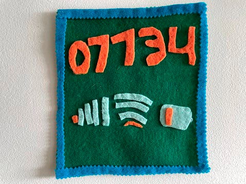 A green felt square with blue trim. Orange felt text reads “07734” or hello, backwards.  Below are 3 felt icons that represent low service, low wifi, low battery. 
