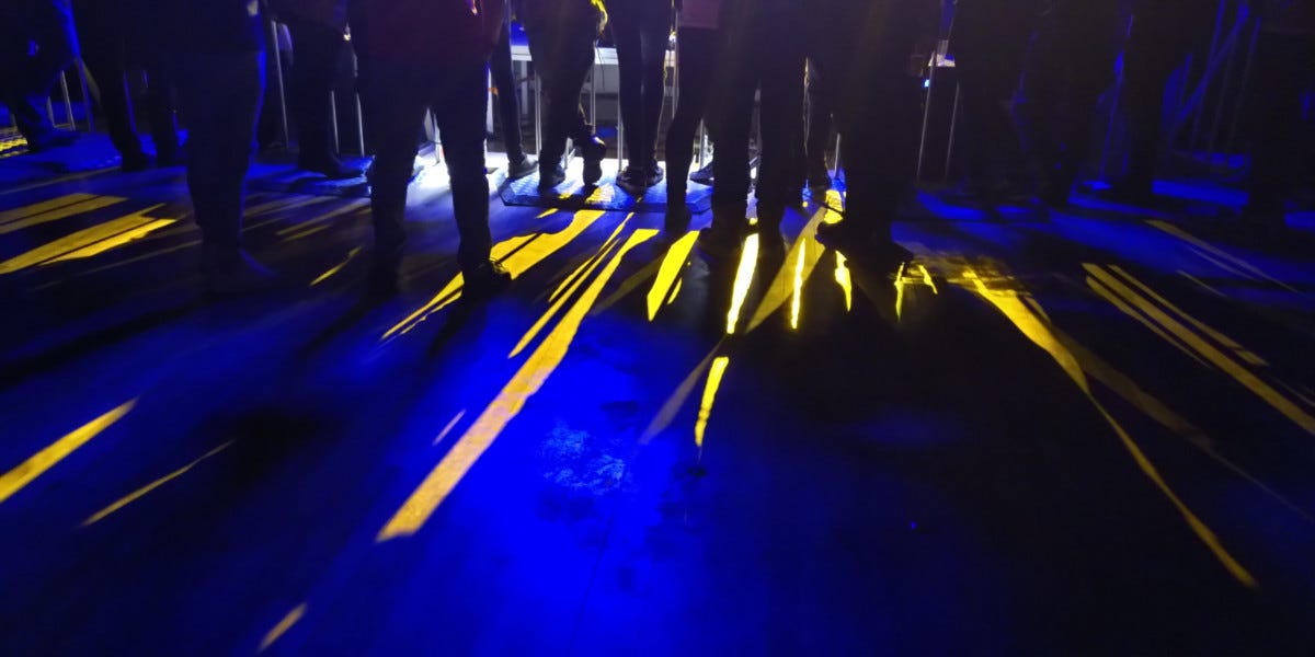 floor lit up by blue and yellow lights coming from a stage, shining on between the silhouettes of legs of people waching the stage