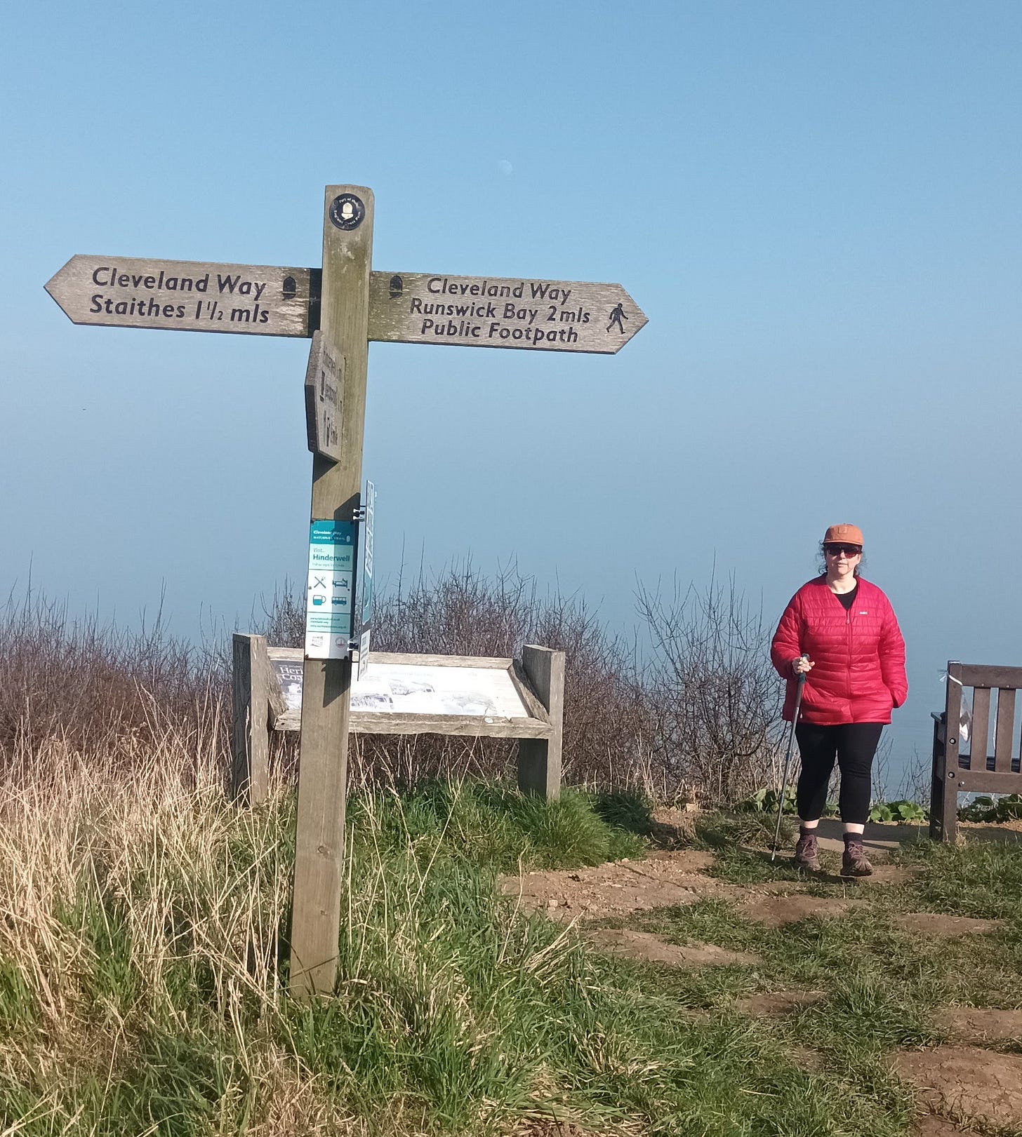 My friend Jo, a white woman in a red coat and orange hat standing next to a footpath sign that says "Cleveland Way, Staithes 1 1/2 mls, Runswick Bay 2mls