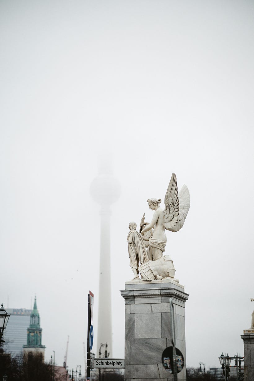 A pale white marble statue of an angel, with a smaller figure in front of her, stands on a grey plinth in a Berlin square. In the background, the Berlin TV tower half-disappears into fog. The top half of the image is dominated by the grey-white sky.