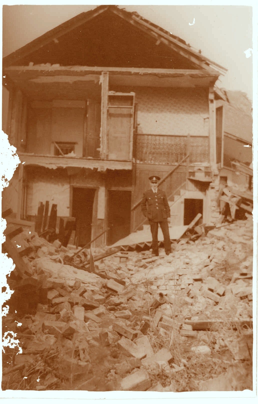A sepia toned photograph of a 2 storey house with the entire front half reduced to rubble. The back wall, roof and back few feet of the rooms are all that remains. A police officer stands on the rubble in the front.
