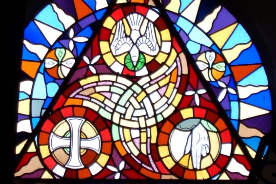 A stained glass image of the Trinity, including a cross, hand, and dove inside a larger triangle.