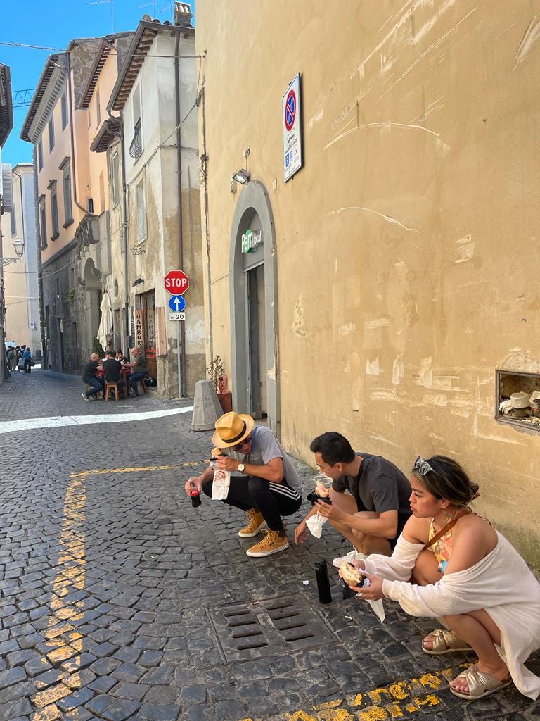 Two men and one woman squatting while eating paninis on a cobblestone street in Orvieto, Italy