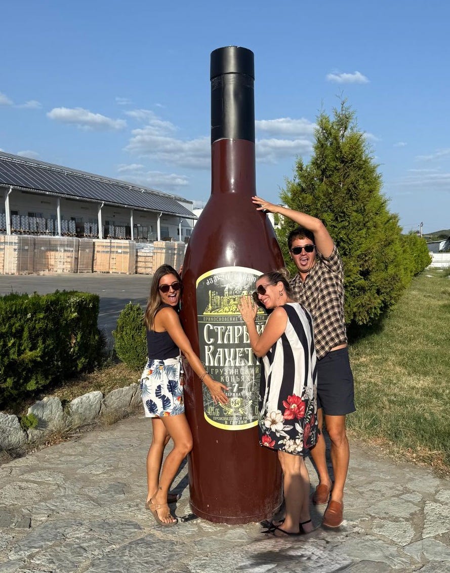 Two white women and one Asian man are hugging a giant wine bottle. It is sunny outside and they are having fun.  