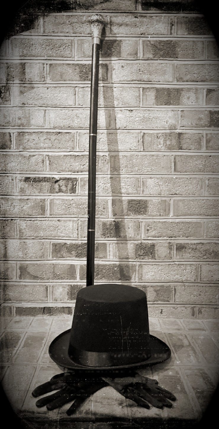 A sepia-toned photo made to look like a Victorian era daguerreotype. The photo shows a black cane with a silver handle leaning against a brick wall with a black top hat and a pair of black gloves sitting on the brick floor in front of the cane.