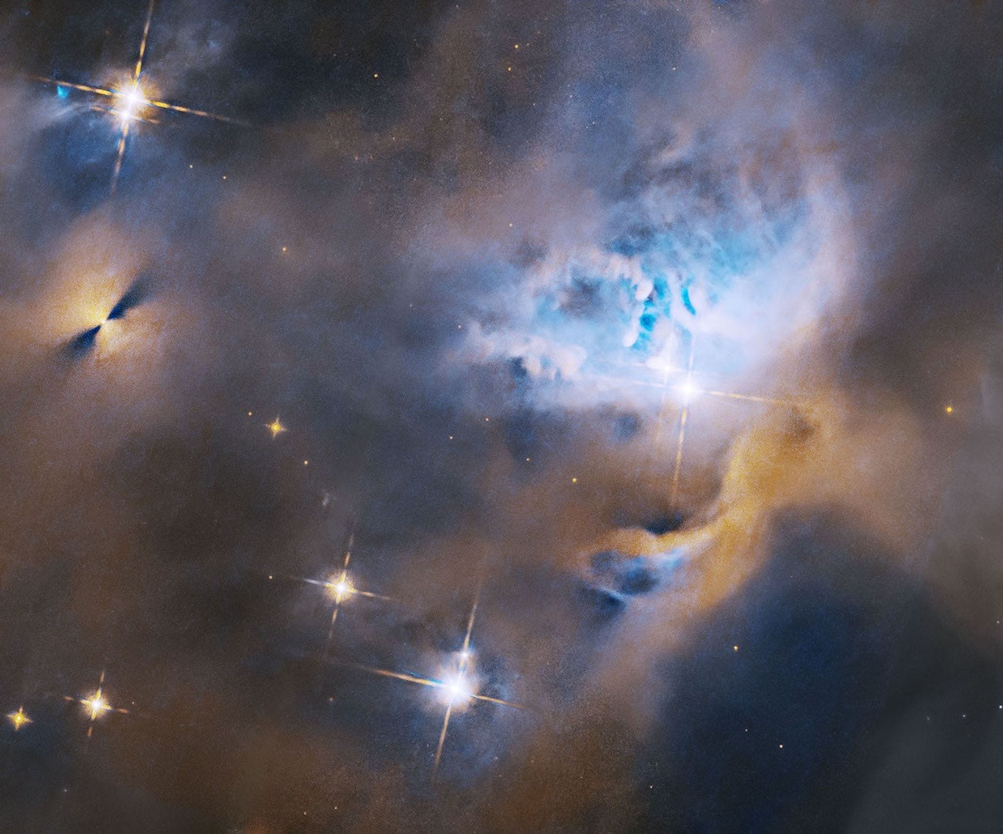 Within a ghostly blue and orange cloud of gas and dust, several stars shine brightly. To the upper left is a protoplanetary disk, appearing as a bright point flanked on either side by fan-shaped shadows. To the center right, stars are nestled in a cavity in the gas and dust. Within a ghostly blue and orange cloud of gas and dust, several stars shine brightly. To the upper left is a protoplanetary disk, appearing as a bright point flanked on either side by fan-shaped shadows. To the center right, stars are nestled in a cavity in the gas and dust.