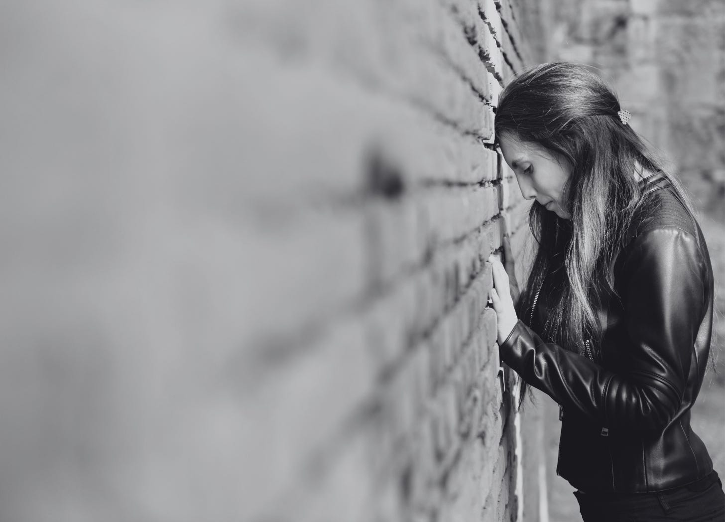 Black and white photo of a woman leaning her forehead against a brick wall, eyes closed, with a solemn expression. Her posture conveys emotional exhaustion or grief, symbolizing quiet suffering and internal struggle.
