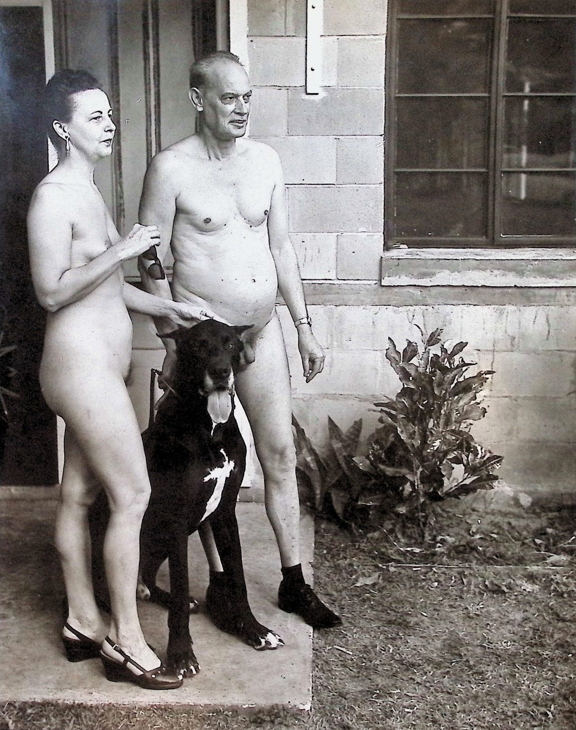 Black-and-white photograph of a nude older couple standing on a concrete walkway in front of a cinder-block building with a large window. The woman, at left, wears wedge sandals and holds the leash of a large black Great Dane sitting at the man's feet; the balding man stands at center wearing only dark shoes.