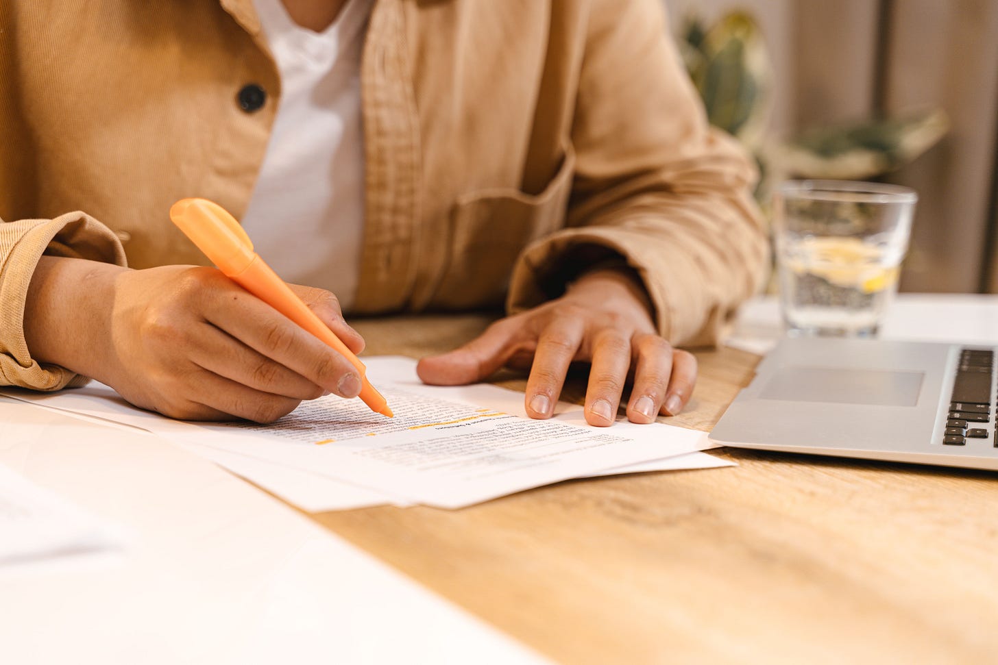 Person highlighting notes at a desk with a laptop and glass of water nearby. Person highlighting notes at a desk with a laptop and glass of water nearby.