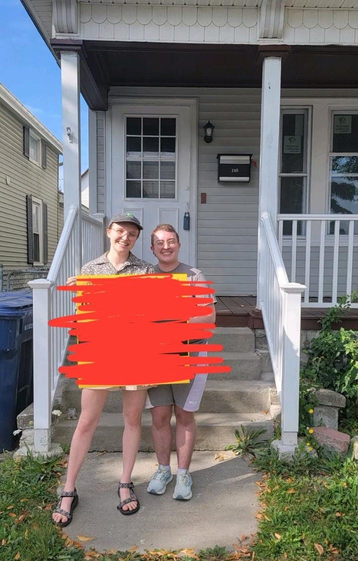 Jo and Emory stand outside of their home, holding a sold sign