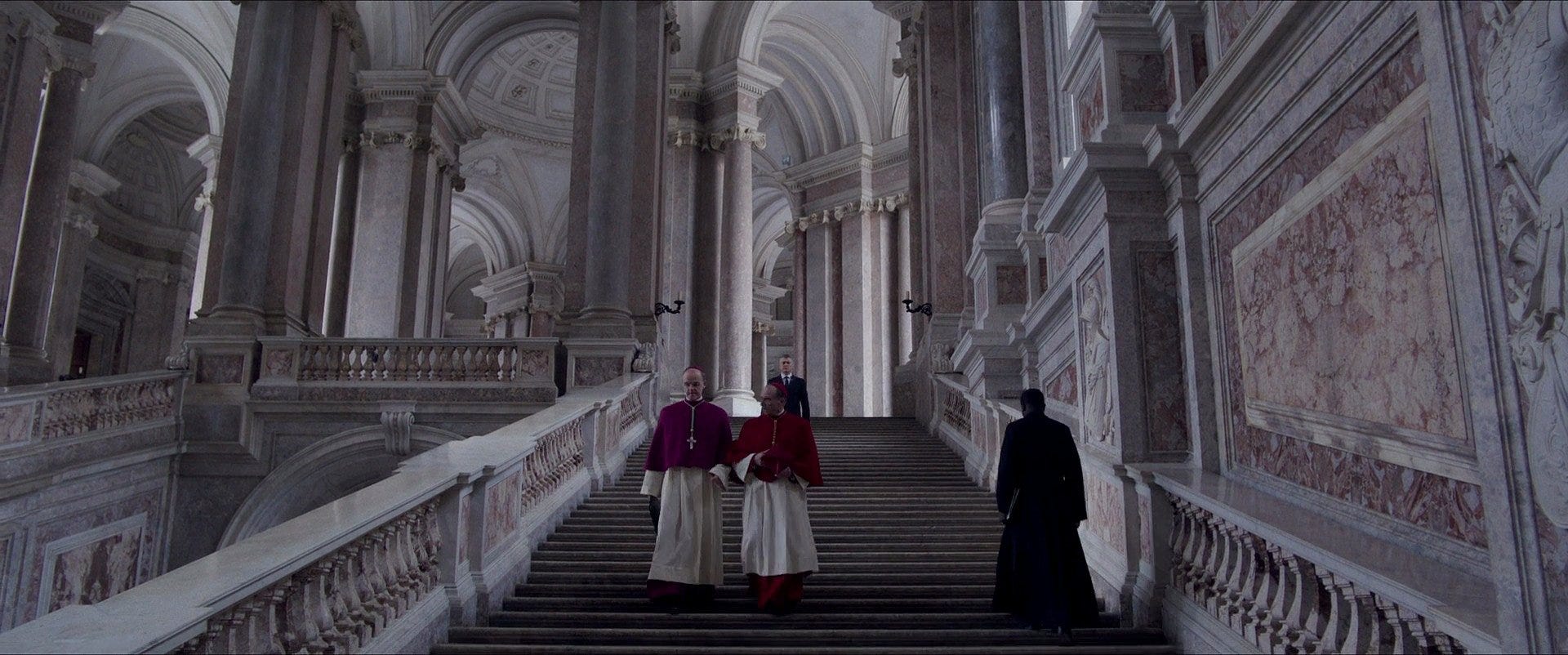 Screenshot from the film "Conclave." Lawrence and Tremblay walk down the stairs. A dark figure walks up.