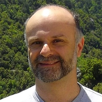 Homem branco de barba grisalha vestindo uma camiseta cinza e sorrindo para a câmera, centralizado na imagem e em frente a um fundo montanhoso coberto por árvores. Homem branco de barba grisalha vestindo uma camiseta cinza e sorrindo para a câmera, centralizado na imagem e em frente a um fundo montanhoso coberto por árvores.