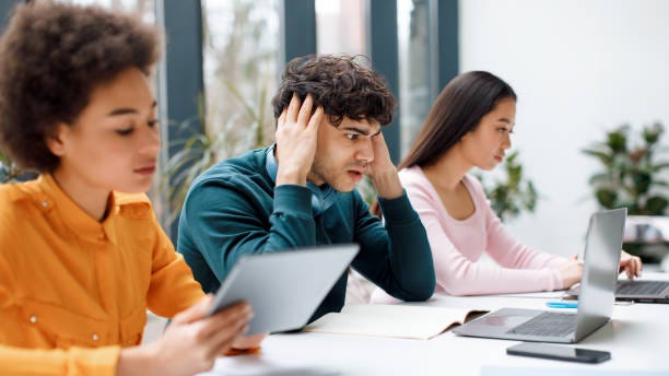 Worried sad student guy looking at laptop, getting final test results, sitting with groupmates in audience