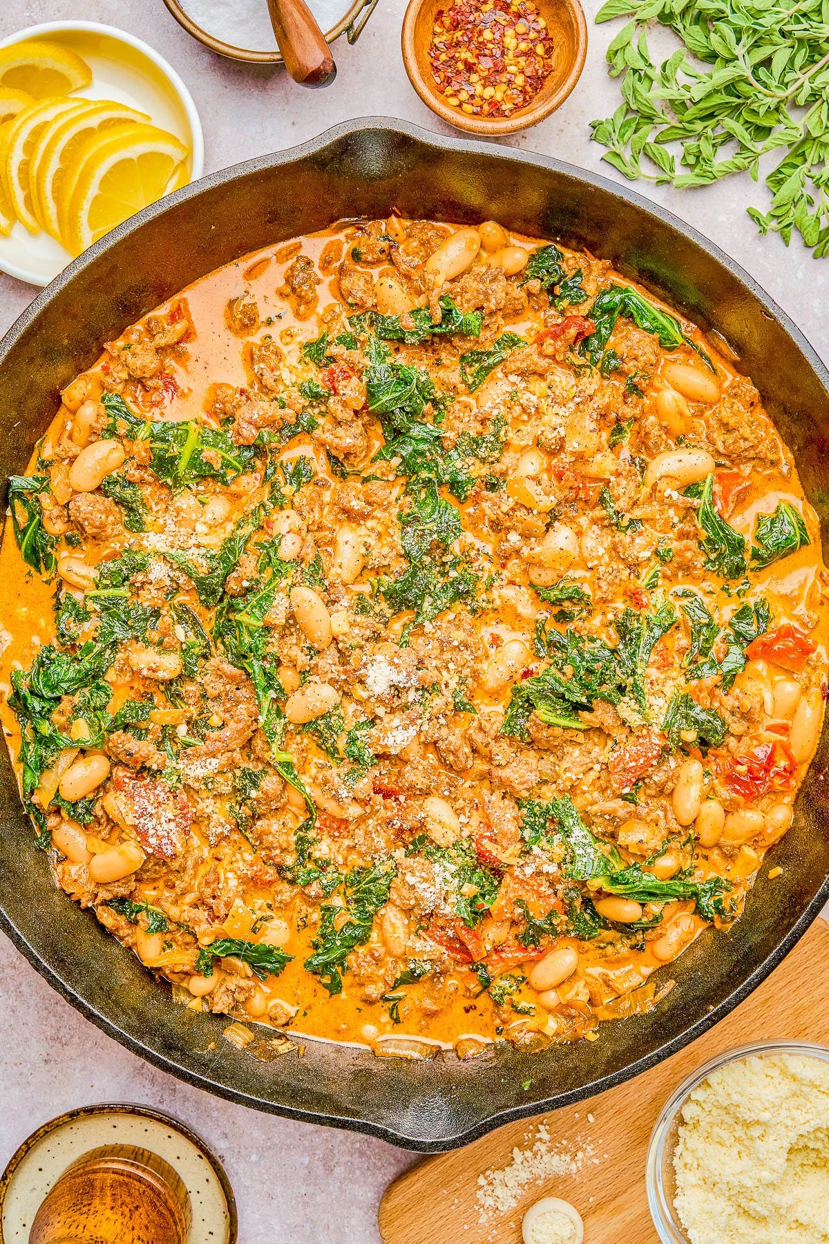 A skillet dish of ground meat, white beans, kale, and tomatoes in a creamy sauce, garnished with grated cheese, surrounded by lemon slices, red pepper flakes, and fresh herbs.