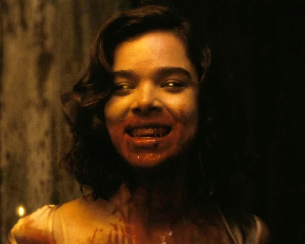 close do busto de uma mulher branca com um sorriso maníaco, olhos que brilham na luz e com a boca e o pescoço completamente ensanguentado. A atriz é Hailee Steinfield, uma mulher branco com cabelos curtos e escuros