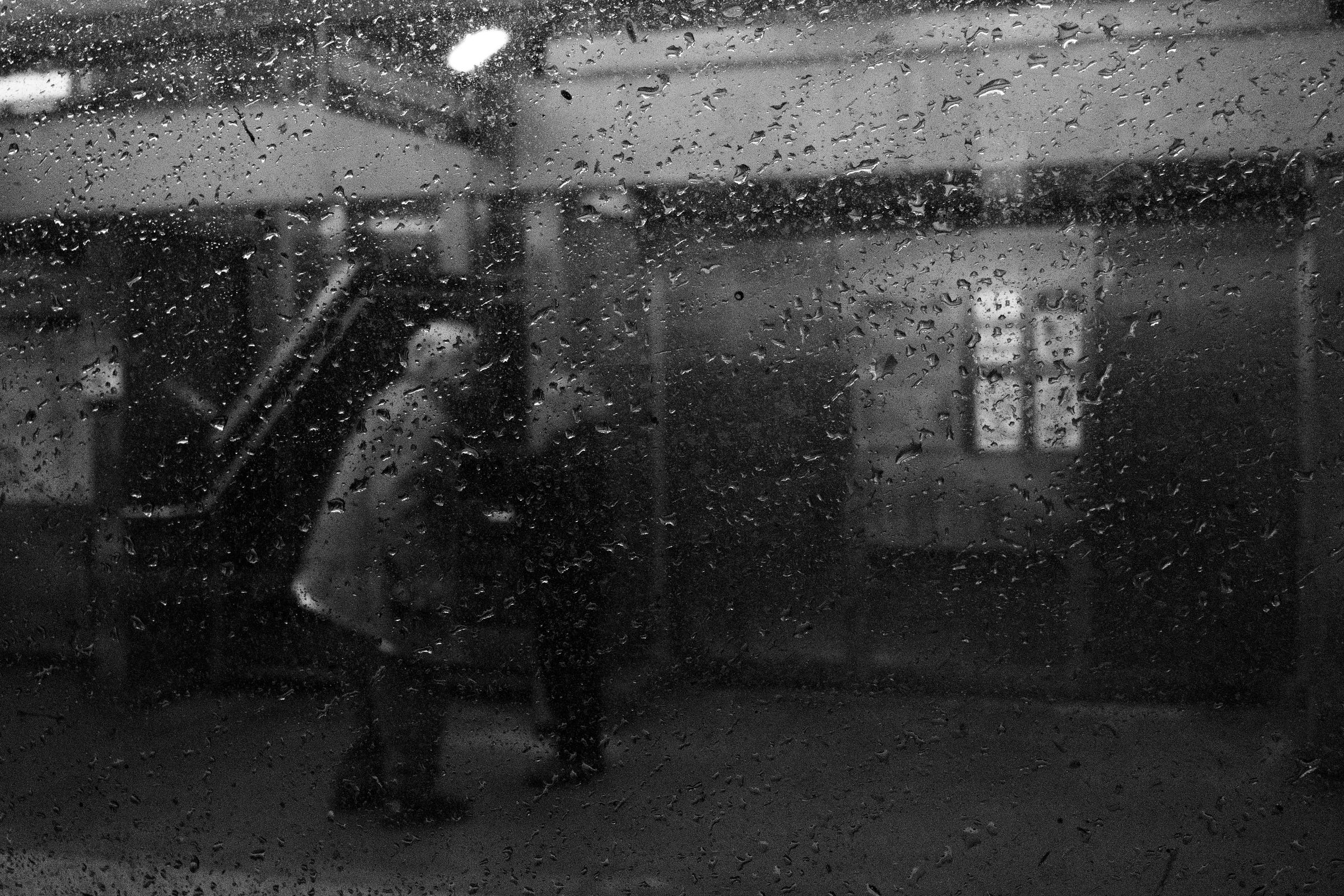 Rain-covered window blurring the view of two people conversing near a staircase at a train station, with overhead lights casting a soft glow. Rain-covered window blurring the view of two people conversing near a staircase at a train station, with overhead lights casting a soft glow.