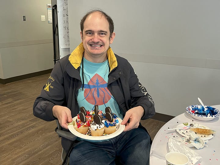James is a middle-aged man who holds a plate of decorated cupcakes. Taylor is a young woman with long hair and glasses who sits beside her plate of decorated cupcakes.