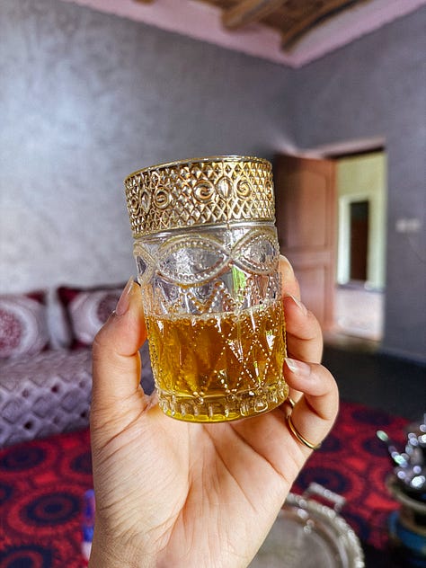 3 photos: 1- Anela holds up a clear embossed glass of tea in one hand, with a gold almost filigree rim. 2-two women ride camels through the desert, looking back smiling. 3-a fire pit glows in the dark in front of a swimming pool and the dark sky above it.