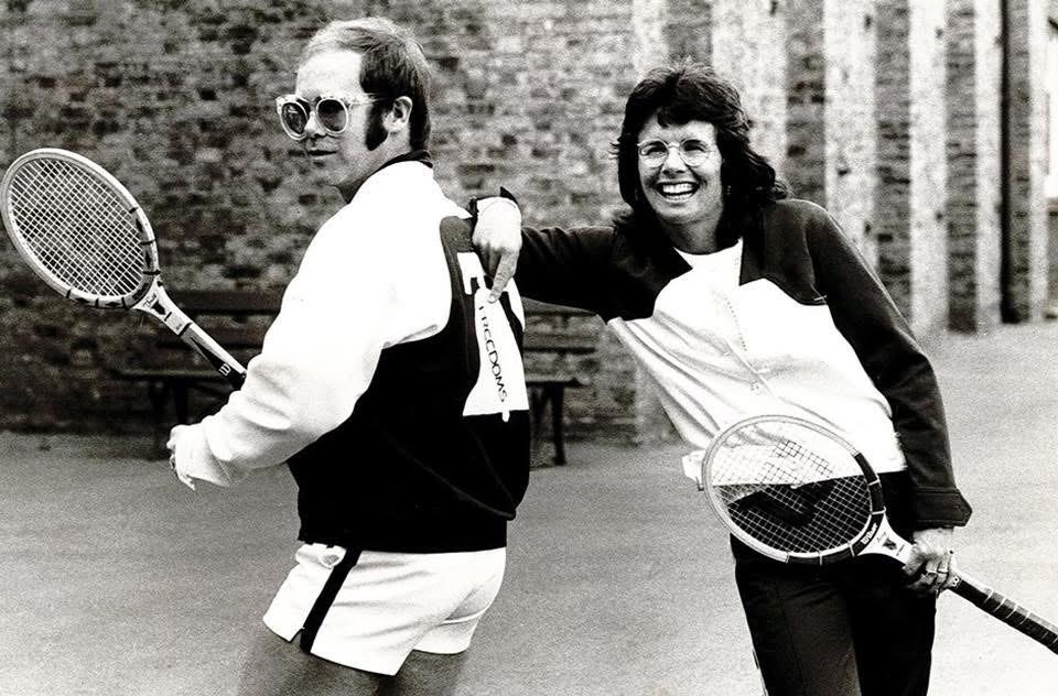 Black and white photo of Elton John and Billie Jean King around 1974. Both are holding tennis racquets. Elton is wearing a Philadelphia Freedoms uniform. Billie Jean is smiling and pointing to the team logo on the back of Elton's jacket.