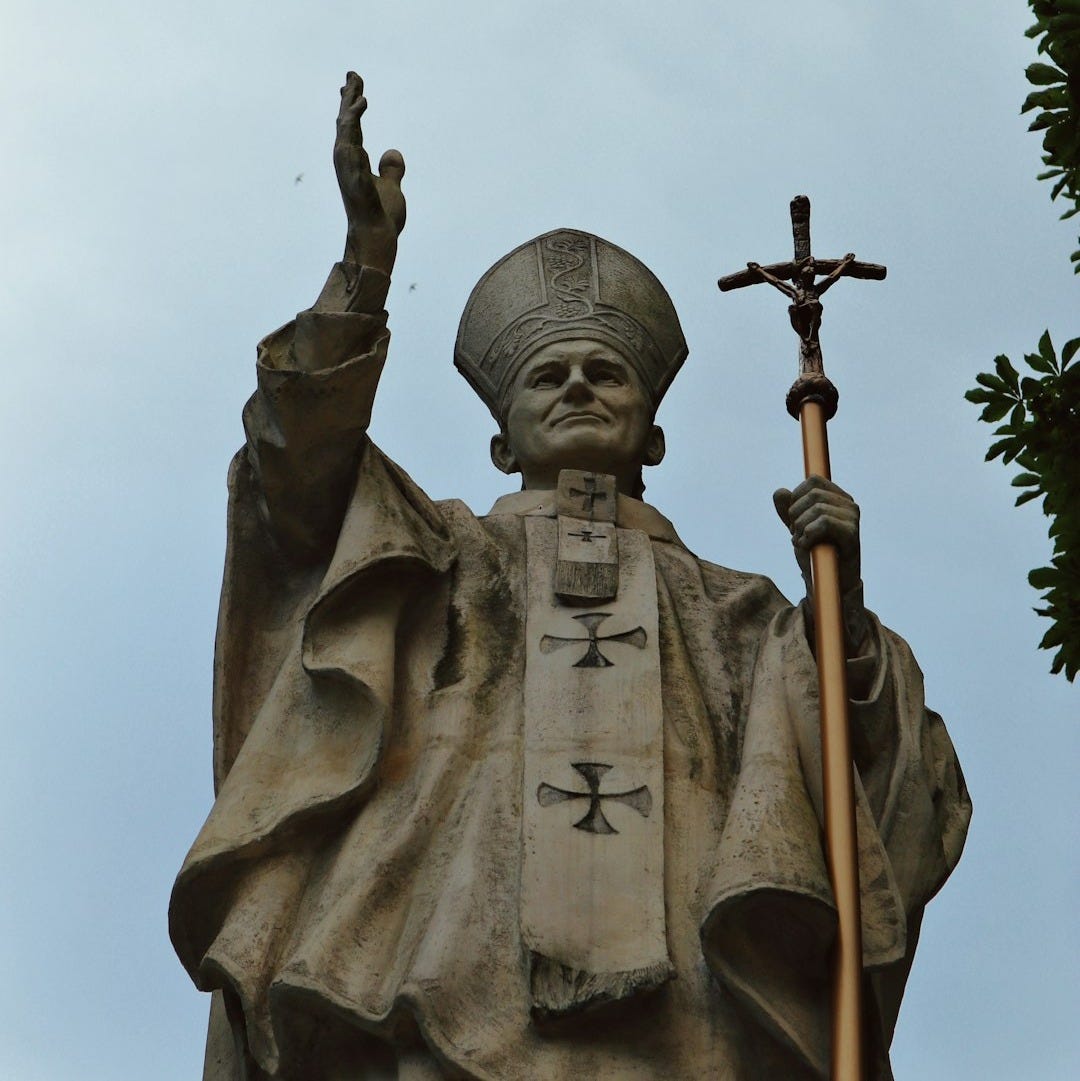 Pope statue with raised hand and crucifix against sky
