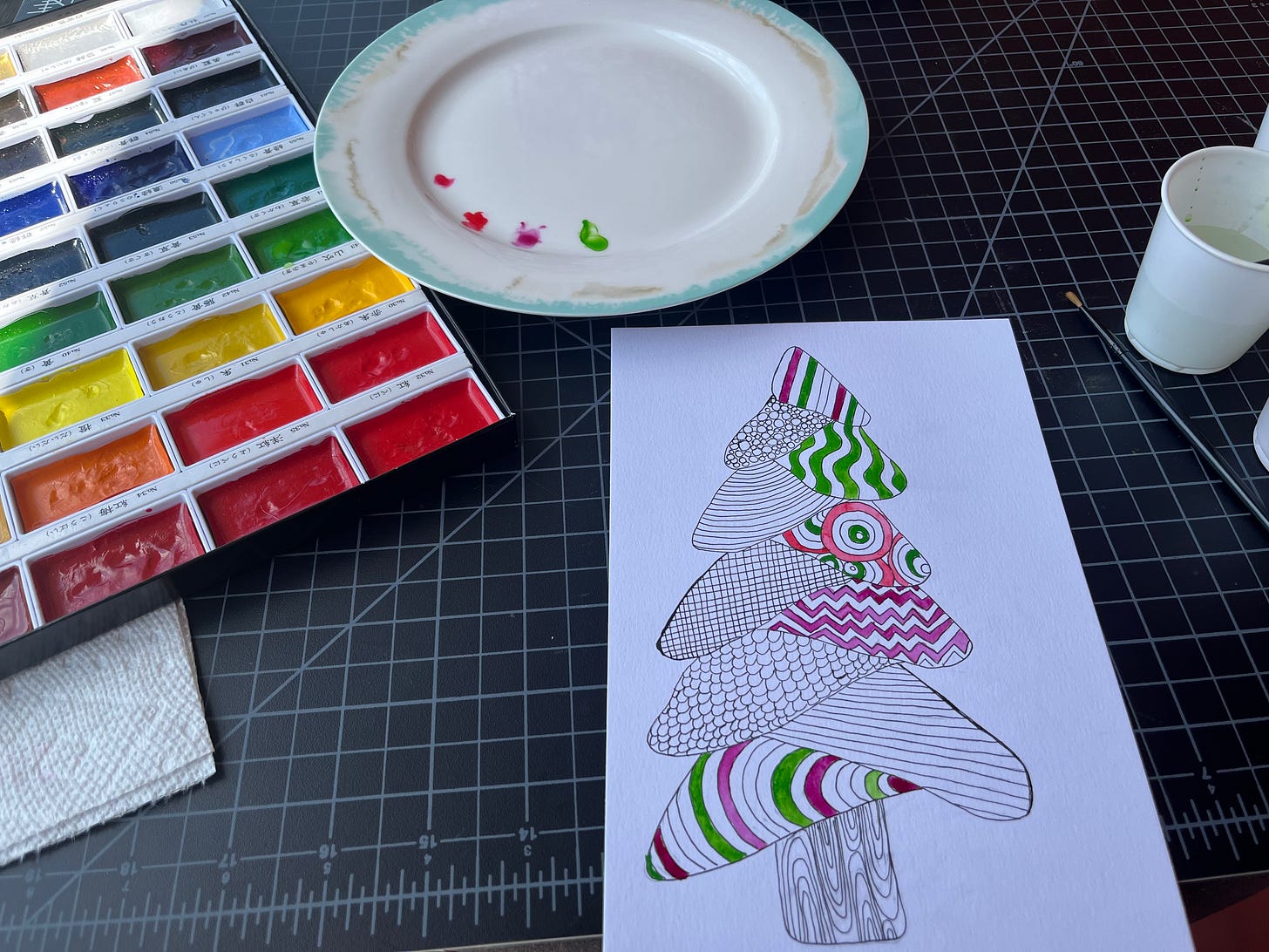 Photo of a partly painted pen and ink illustration of a funky Christmas tree beside watercolor paints and palette.