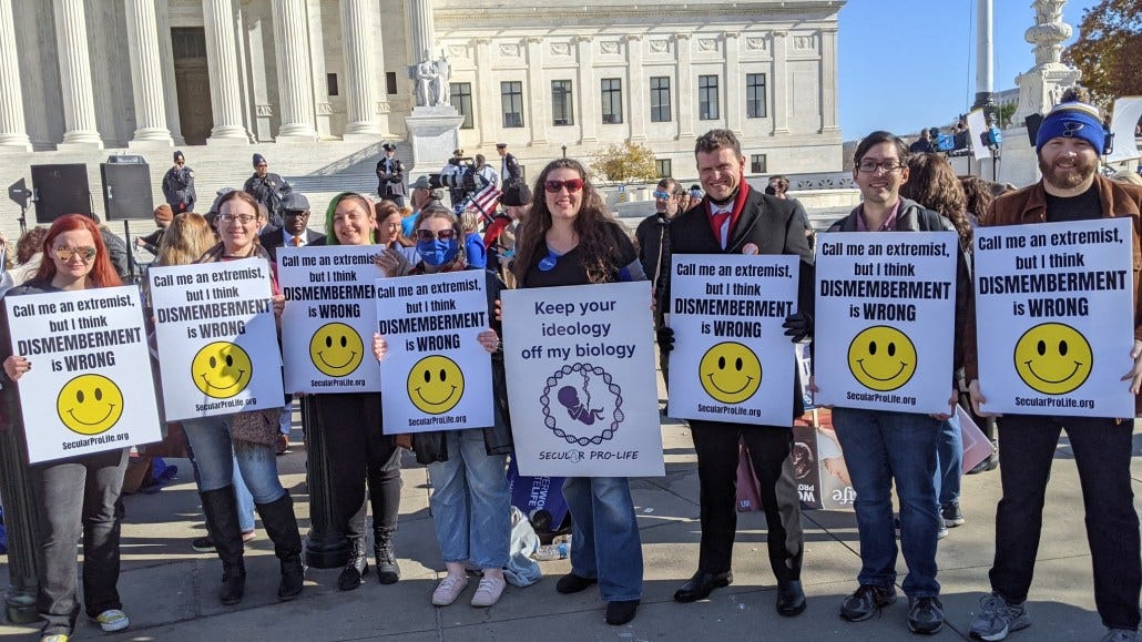 Secular pro-lifers stand outside the Supreme Court building. They hold signs reading "Keep your ideology off of my biology" and "Call me an extremist, but I think dismemberment is wrong."