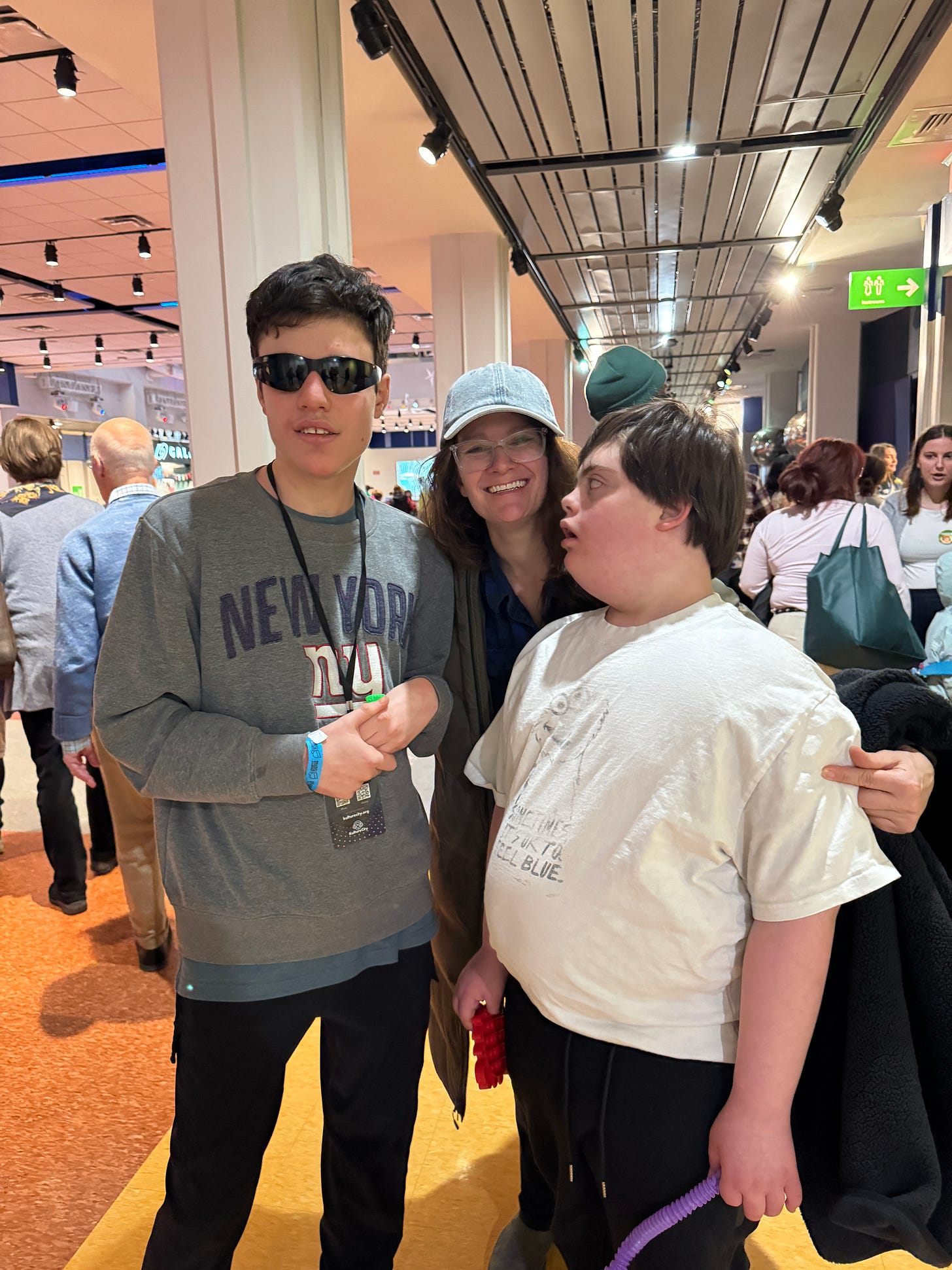 Two young men stand beside one another. The young man on the left wears sunglasses and a NY Giants sweatshirt, and has short dark brown hair. The young man on the right is facing him, looking up at him, wearing a white t-shirt and holding two sensory fidgets. Between them, a middle-aged white woman wearing a denim baseball hat smiles. Two young men stand beside one another. The young man on the left wears sunglasses and a NY Giants sweatshirt, and has short dark brown hair. The young man on the right is facing him, looking up at him, wearing a white t-shirt and holding two sensory fidgets. Between them, a middle-aged white woman wearing a denim baseball hat smiles.
