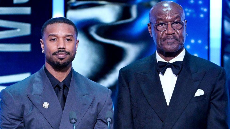 Michael B. Jordan wears a navy blue perfectly tailored suit with a black collar shirt and matcing tie, adorned with a white brooch. Delroy Lindo wears and black perfectly tailored tuxedo complete with white pocket square. Both powerful and accomplished Black men standing on the BAFTAs stage to present an award seemlingly frown in a polite disgust of being called the N-word by John Davidson, a white man with Tourette Syndrome who was in the audience. Image from Getty Images. Michael B. Jordan wears a navy blue perfectly tailored suit with a black collar shirt and matcing tie, adorned with a white brooch. Delroy Lindo wears and black perfectly tailored tuxedo complete with white pocket square. Both powerful and accomplished Black men standing on the BAFTAs stage to present an award seemlingly frown in a polite disgust of being called the N-word by John Davidson, a white man with Tourette Syndrome who was in the audience. Image from Getty Images.