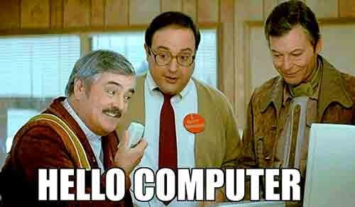 Scene from Star Trek IV: The Voyage Home showing three men gathered around a computer. The man on the left, wearing a red and yellow outfit, humorously speaks into a computer mouse. The middle man, in glasses and a beige sweater with a red button, and the man on the right, in a brown jacket, watch the screen with interest. Bold white text at the bottom reads “HELLO COMPUTER,” highlighting the comedic clash between futuristic expectations and 20th-century technology.