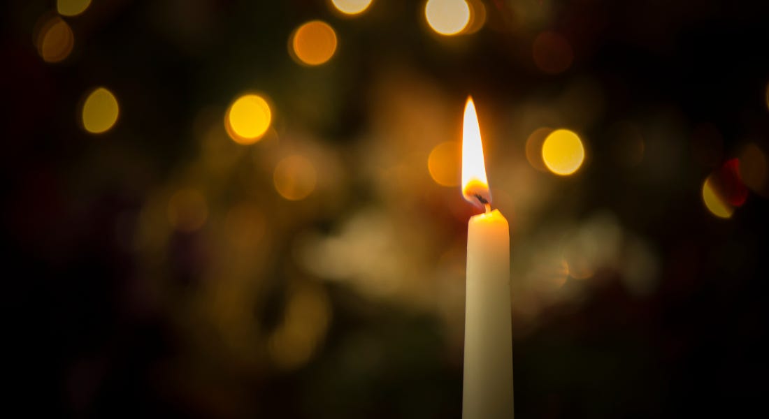 A tall white candle with a flame and twinkling lights behind it