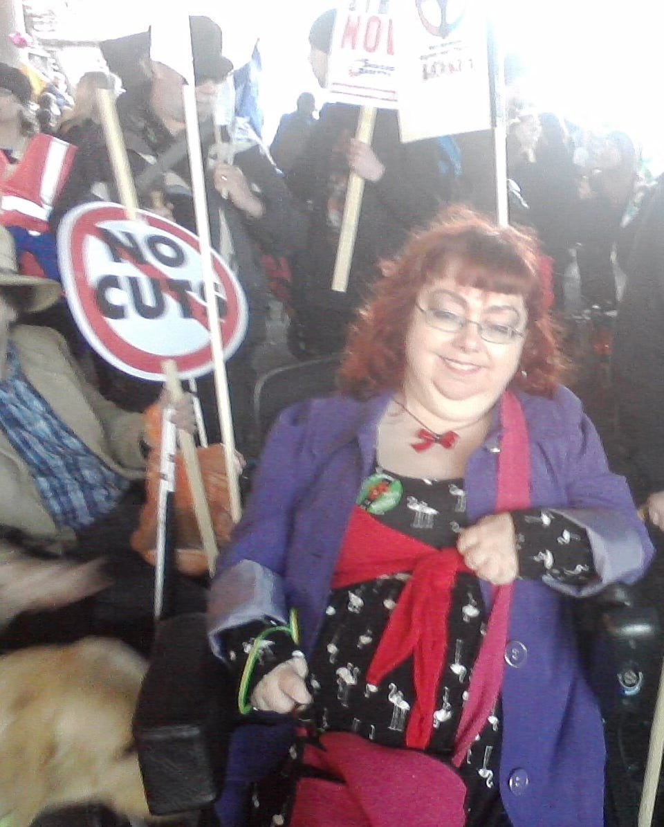 Penny is in her wheelchair in a crowd of people cramped together. She is at the front wearing a purple mac, a black and white dress and a red cardigan. She wears glasses and has red hair. To her left is a man in a brown jacket holding up a sign saying no cuts. It is a typical no entry graphic, a red circle on white with a line through it. There are many posters held up by countless disabled people. The image has a misty quality and was taken under Waterloo Bridge. The vibe is intense but upbeat for a mass protest.