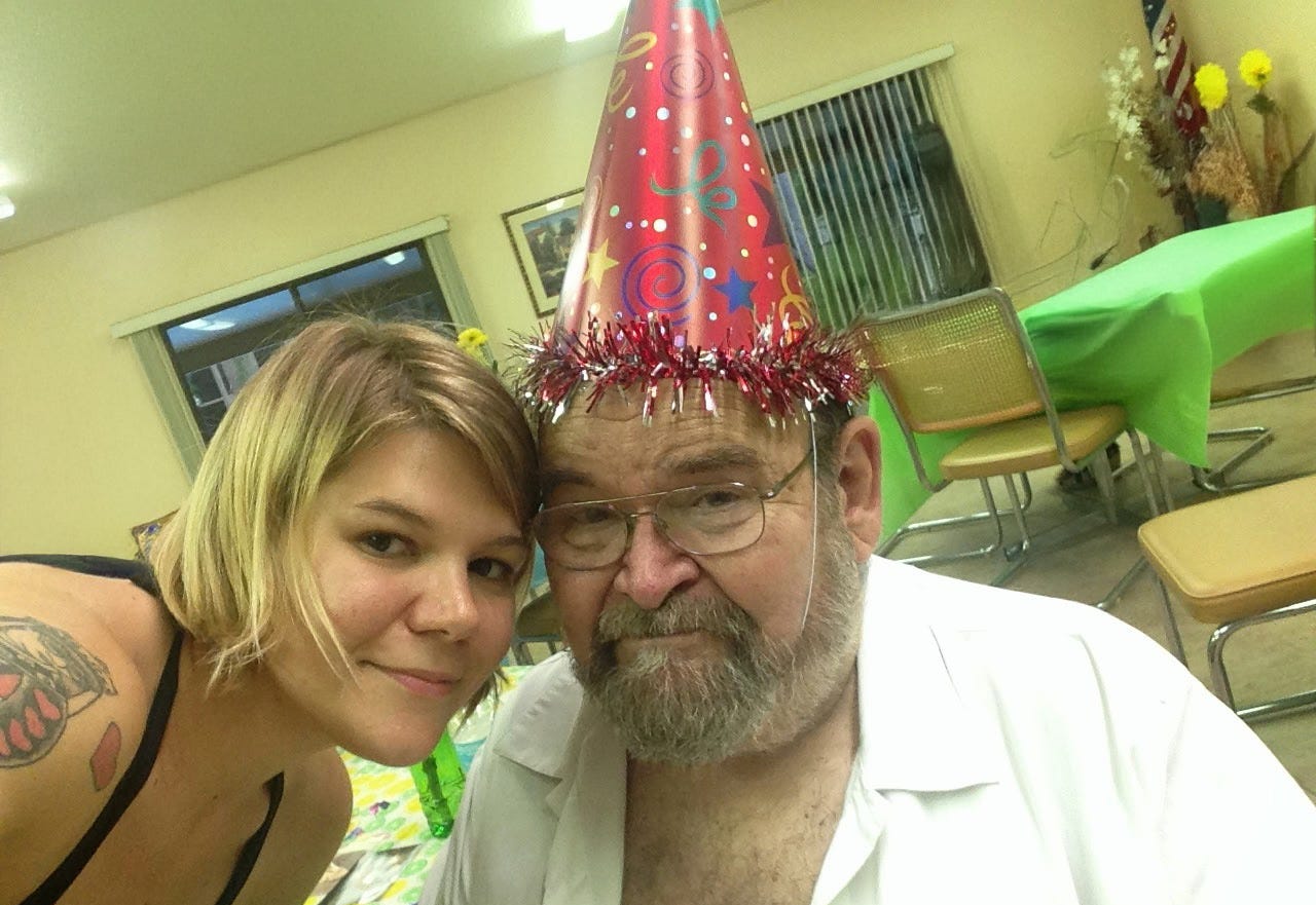 A youngish woman smiles next to her elderly father, who is wearing a red party hat and smiling.