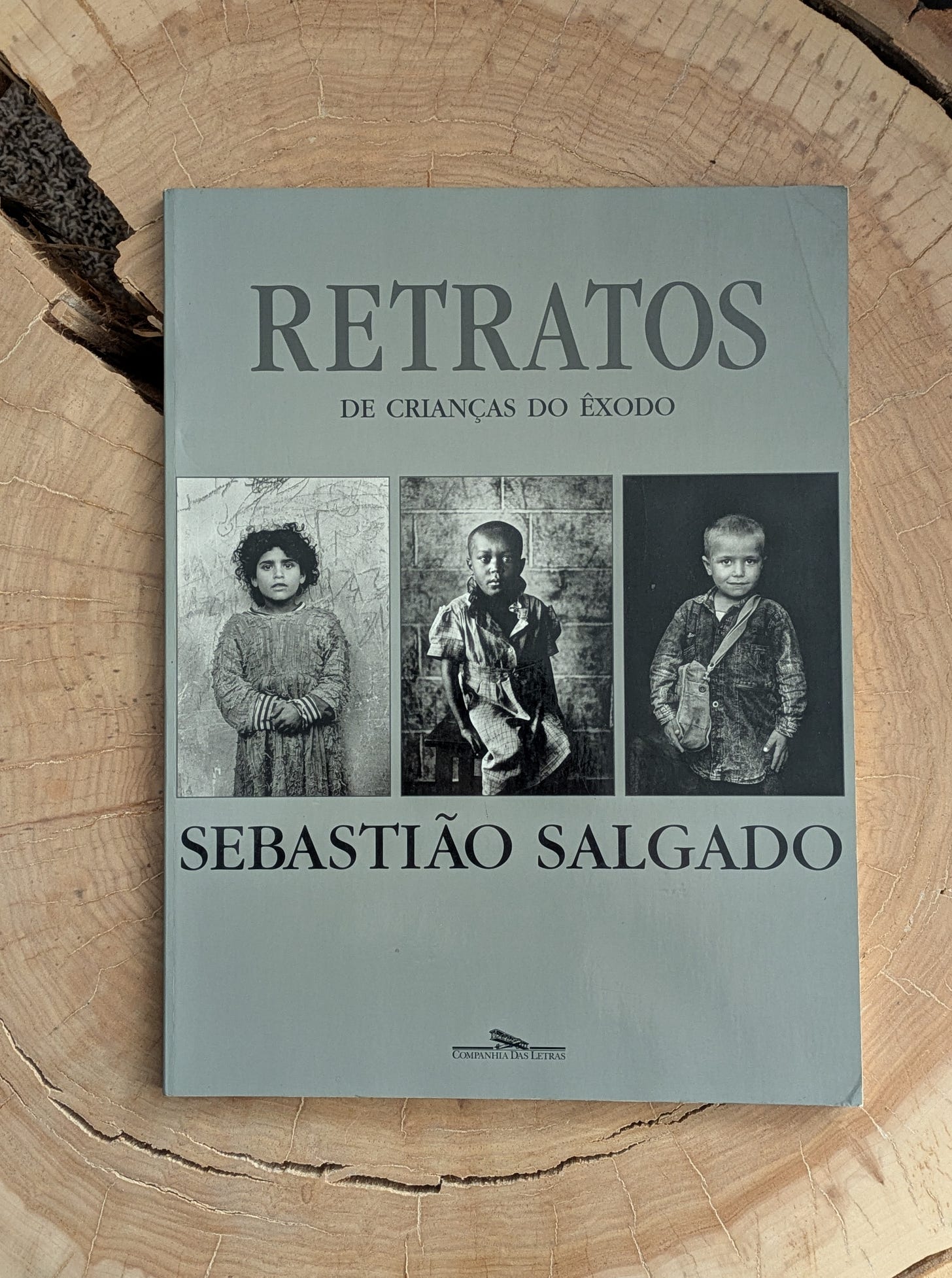 Photo of the book by Sebastião Salgado titled Retratos de Crianças do Êxodo. The book cover shows three black and white photos of migrant children. The book is sitting on a cracked tree stump.