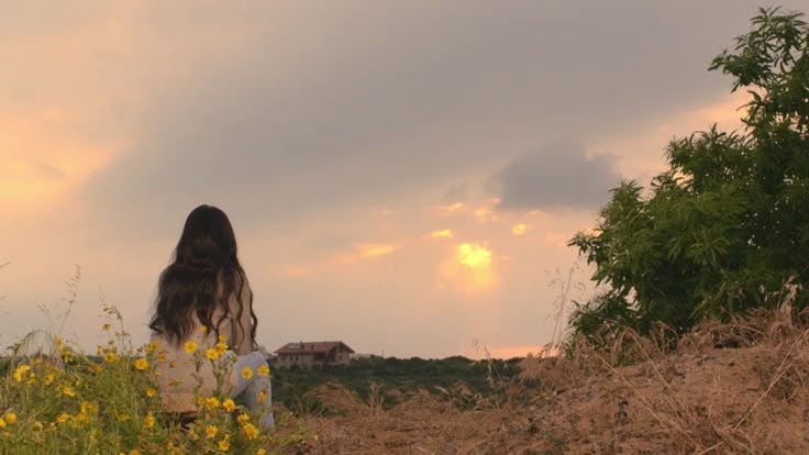 This may contain: a woman sitting on top of a field next to a tree and yellow wildflowers