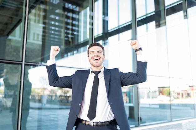 Cheerful Hispanic businessman raising arms and celebrating victory outside office building