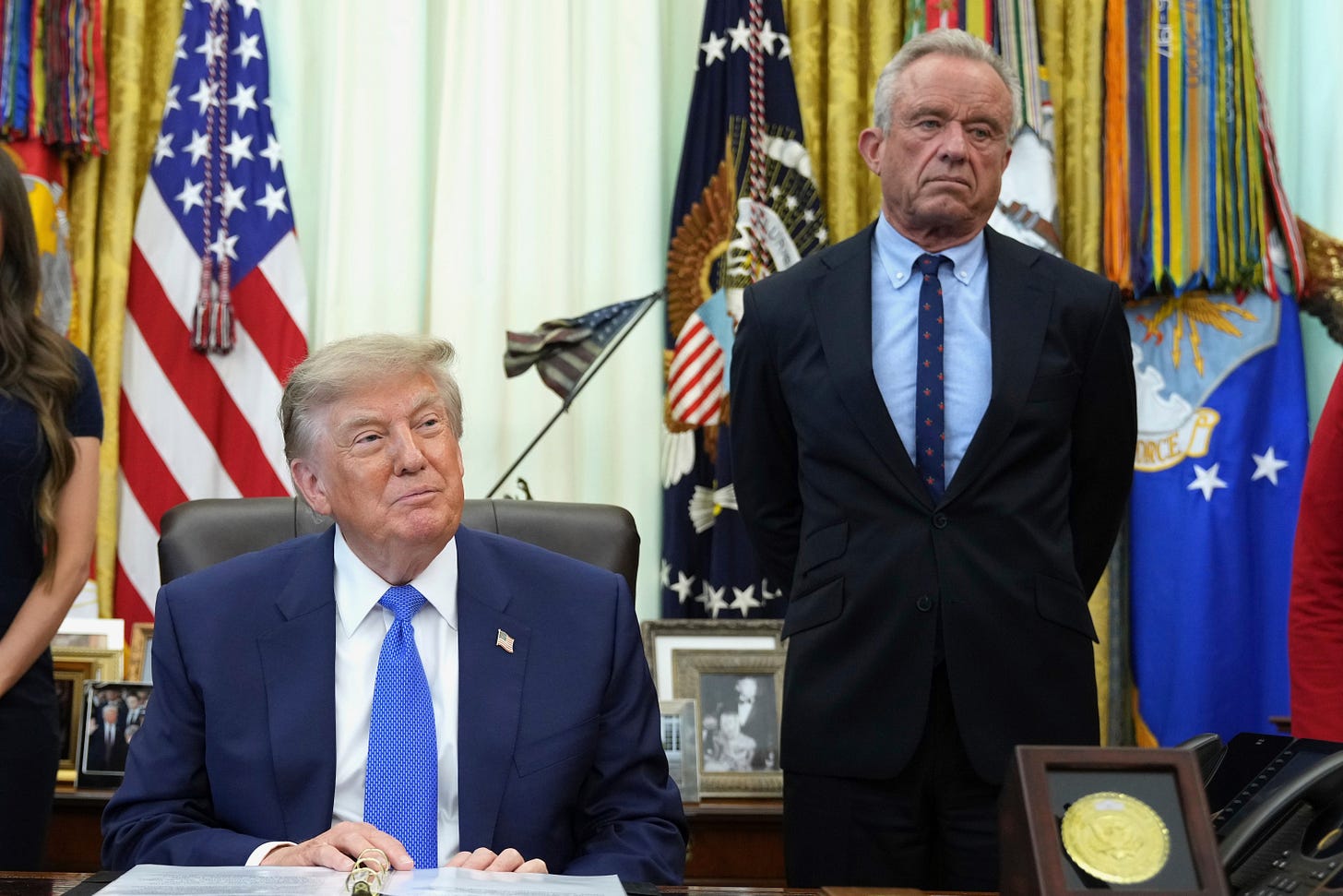 President Trump sits at his desk in the Oval Office with RFK Jr. standing beside him near military flags. President Trump sits at his desk in the Oval Office with RFK Jr. standing beside him near military flags.