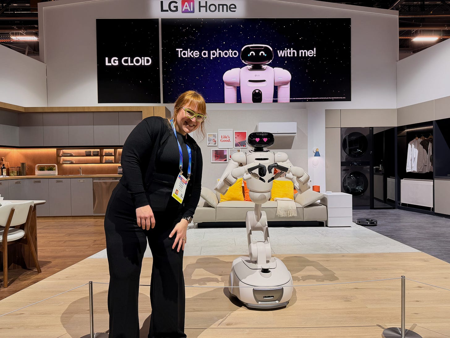 Tiffany Kyazze poses next to LG’s CLOiD household robot at CES, standing in a staged smart home setup with a large “Take a photo with me!” display behind them. The robot is making a heart gesture with its hands. Tiffany Kyazze poses next to LG’s CLOiD household robot at CES, standing in a staged smart home setup with a large “Take a photo with me!” display behind them. The robot is making a heart gesture with its hands.