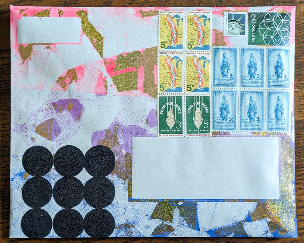 A handmade envelope with a splattered pink, gold, and white background is adorned with an eclectic mix of vintage U.S. postage stamps. Black circular stickers form a pattern in the lower left corner, while blank address fields remain open for mailing.