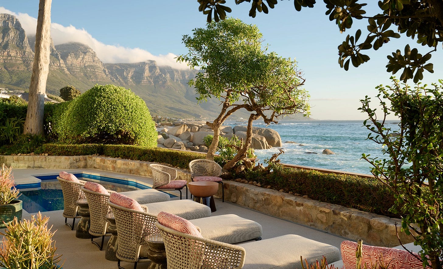Perfect Hideaways luxurious oceanfront terrace with woven loungers and patterned cushions beside a private plunge pool, surrounded by sculpted greenery and overlooking boulders and the turquoise Atlantic Ocean with the Twelve Apostles mountain range in the background, framed by trees under a golden sunset glow, Camps Bay, Nalu Villa