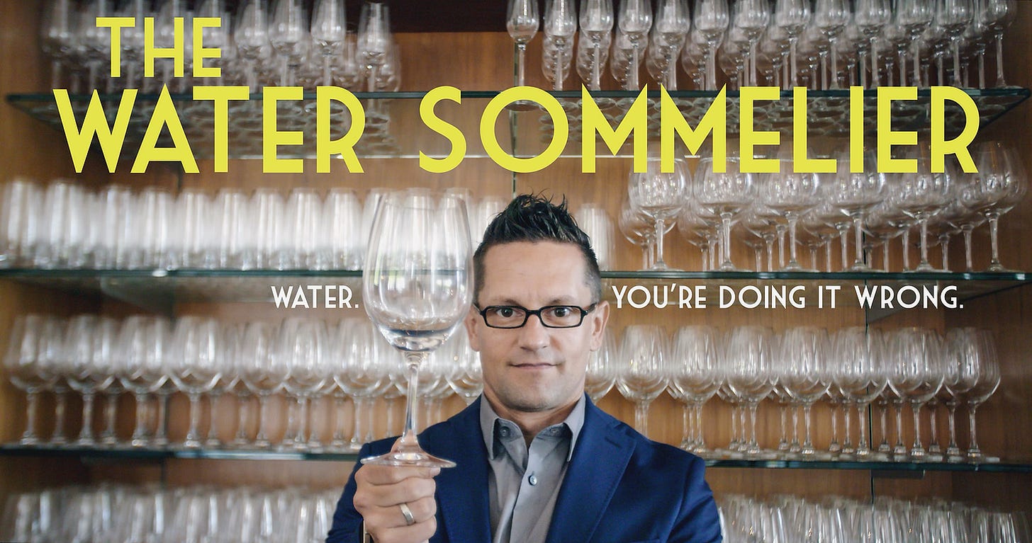 Man in a blue suit holding a wine glass, standing before shelves of glasses. Text: "The Water Sommelier. Water. You're doing it wrong."