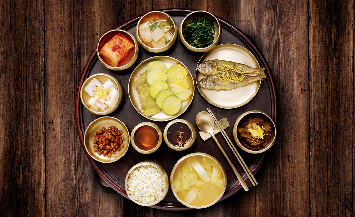 Overhead photo of a traditional Korean meal arranged on a large round dark tray placed on a wooden table. The setting includes a bowl of white rice, a pale soup, grilled fish, kimchi, seasoned greens, pickled vegetables, small dishes of sauces, beans, and several vegetable side dishes. A spoon and chopsticks rest near the center-right of the tray. The image emphasizes the structure of a Korean meal as a full spread, with many dishes placed together rather than served one course at a time.
